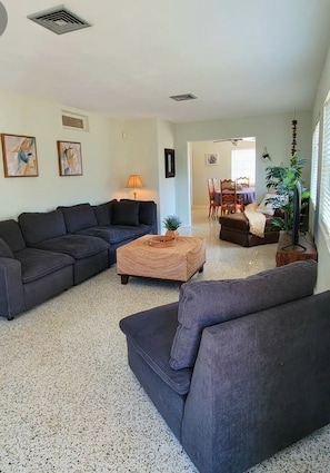 A living room with a sectional sofa, a coffee table, and a television. There is a dining area in the background with a table and chairs.