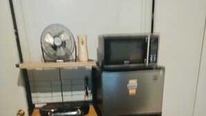 A compact kitchen with a microwave, a small refrigerator, a fan, and a wooden shelf unit. - Perfect 2-bedroom apartment with AC in charming Bronx neighborhood (Bronx)