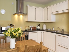 A kitchen with white cabinets, a stainless steel range hood, a wooden dining table with chairs, and a vase of yellow flowers. - 2 Bed in Crickhowell (oc-bn192) (Llangynidr)