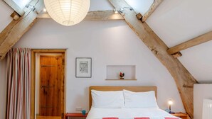 Un dormitorio con techo inclinado, vigas de madera, una cama con ropa de cama blanca y roja, una mesita de noche pequeña, una mesita auxiliar y una estantería en la pared, además de una puerta.