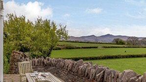 Un muro de piedra con un banco curvo y una mesa, un árbol y vistas a un campo verde y colinas.