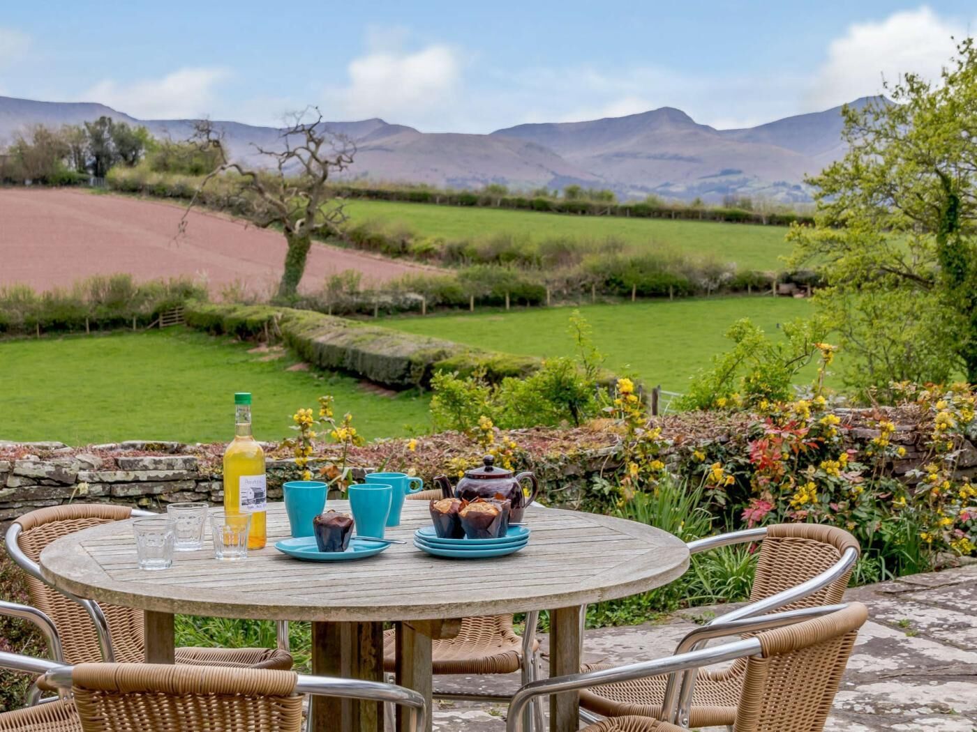 Un área de comedor al aire libre con una mesa para dos, rodeada de sillas de mimbre, con una vista panorámica de colinas y un jardín al fondo.