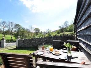 Un espace extérieur aménagé pour les repas, avec un mur en pierre, des bancs en bois et une table dressée pour deux personnes, avec des assiettes, des verres et une carafe de jus d’orange.