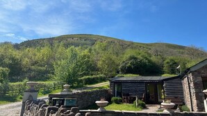 Una cabaña rústica con un muro de piedra, una terraza de madera y vistas a una colina verde.