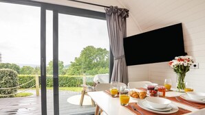 A dining area with a table set for breakfast, a TV mounted on the wall, and a view of greenery outside.