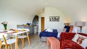 A compact room with a dining table, a bed with a red blanket, a kitchen area, and a cow-themed artwork on the wall.