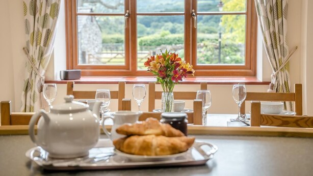 A breakfast setting with a table, tea set, croissants, and a vase of flowers by a window with a view of greenery. - 3 Bed in Brecon (oc-bn370) (Llyswen)