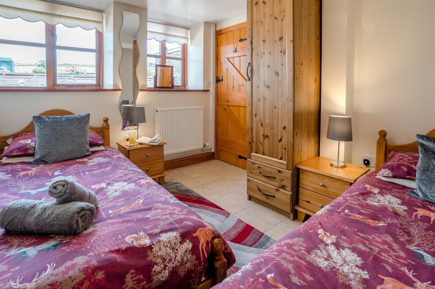 A bedroom with two beds, wooden wardrobe, bedside tables, and a window with a view.