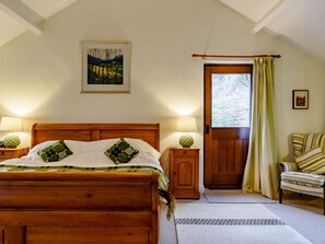 A bedroom with a wooden bed, two bedside lamps, a door with curtains, a chair, and a painting on the wall. - 5 Bed in Crickhowell (oc-bn365) (Llangattock)