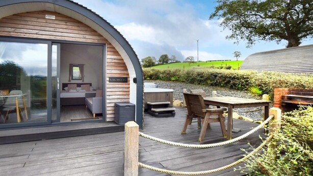 Ein gemütliches, halboffenes Blockhaus mit Glastür, die zu einem möblierten Innenraum führt, einer Terrasse mit Sitzgelegenheit und Blick auf eine grüne Landschaft.