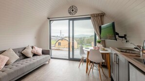 Ein modernes Wohnzimmer mit grauem Sofa, einem Essbereich mit Tisch und Stühlen, einer Küche mit Fernseher und einem großen Fenster mit Blick auf eine ländliche Landschaft.