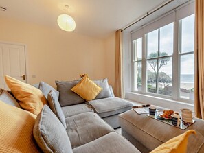 A cozy living room with a sofa set, a coffee table with breakfast items, and a view of the sea through large windows. - 1 Bed in Tenby (oc-fb021) (Tenby)