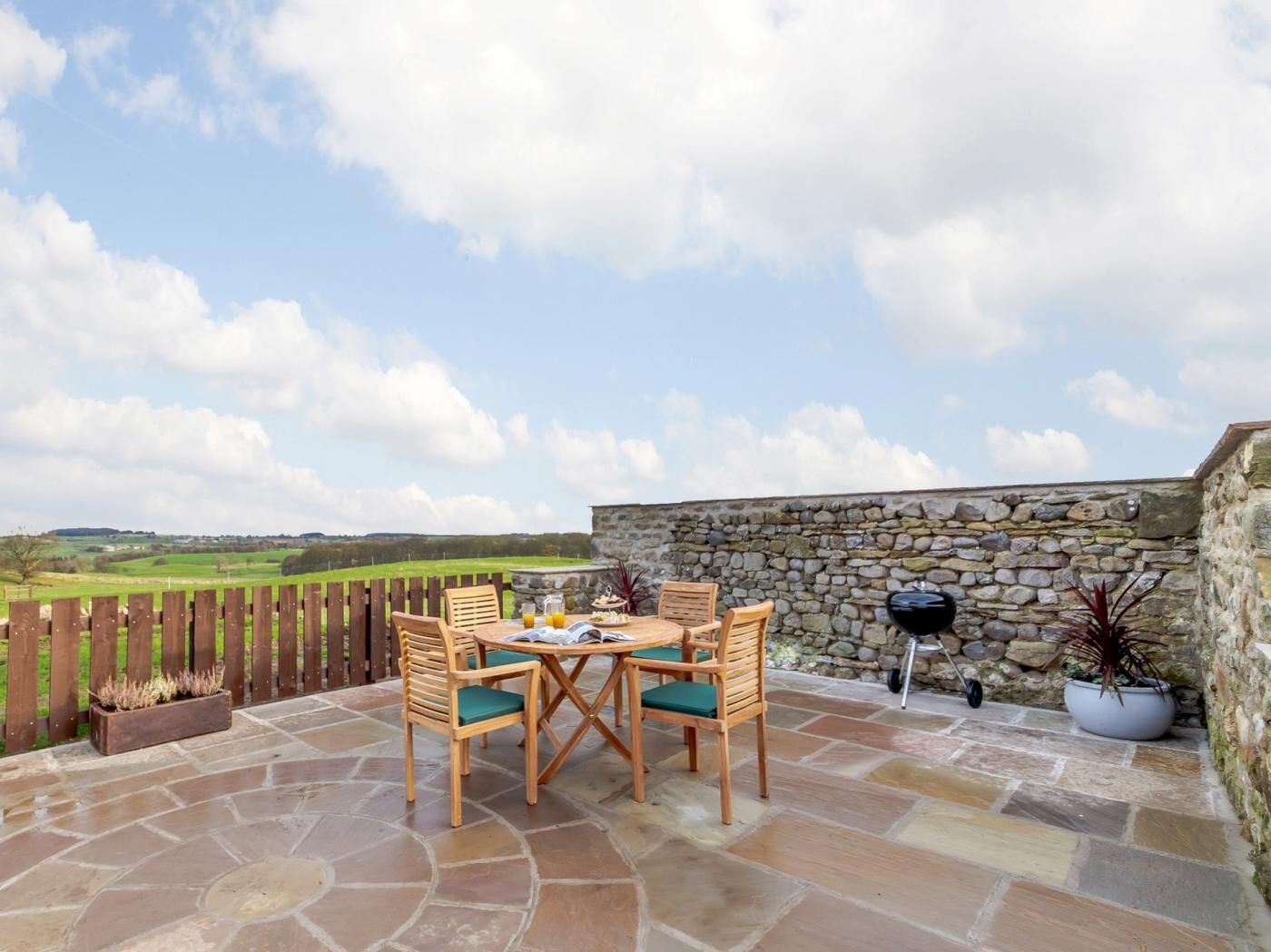 A patio with stone flooring, a wooden table with chairs, a stone wall, and a view of the countryside.
