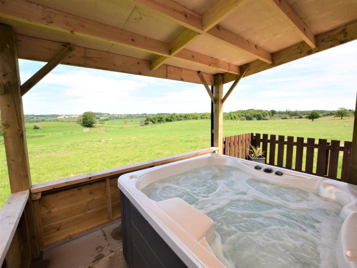 A hot tub under a wooden pergola with a view of a green field.
