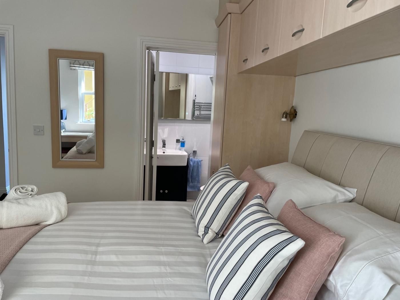 Un dormitorio con cama, almohadas, un espejo, un armario y un baño visible a través de una puerta abierta.