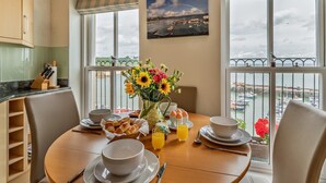 En matplats med ett bord dukat för frukost, en vas med blommor och utsikt över havet genom stora fönster.