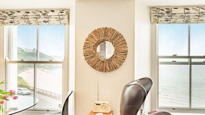 A room with a view of a beach and a body of water, a round mirror, a table with a chair, and a radio on a small wooden side table. - 2 Bed in Tenby (oc-fb110) (Tenby)