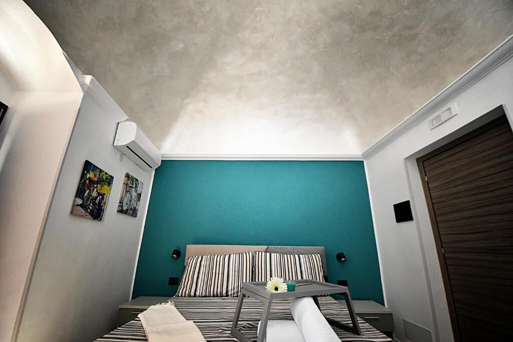 Ein kleiner, kompakter Raum mit einer Türkis-farbenen Akzentwand, einem Bett mit gestreifter Bettwäsche, einem kleinen Tisch mit einer Blume und einem Deckenventilator.