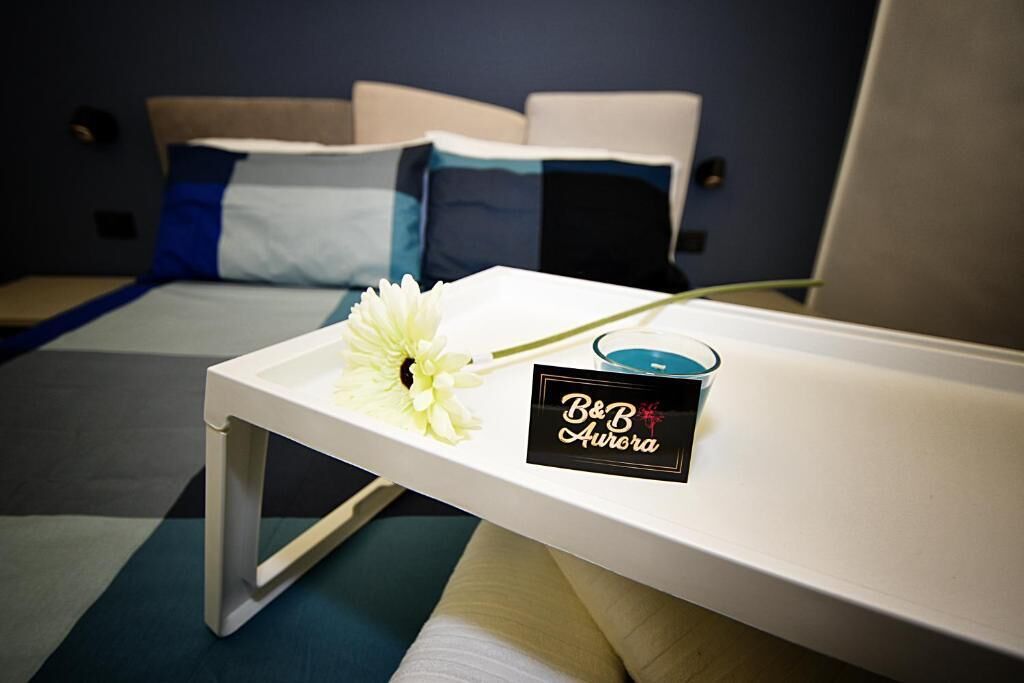 A hotel room with a bed, a bedside table with a flower and a candle, and a sign reading 'B&B Aurora'.