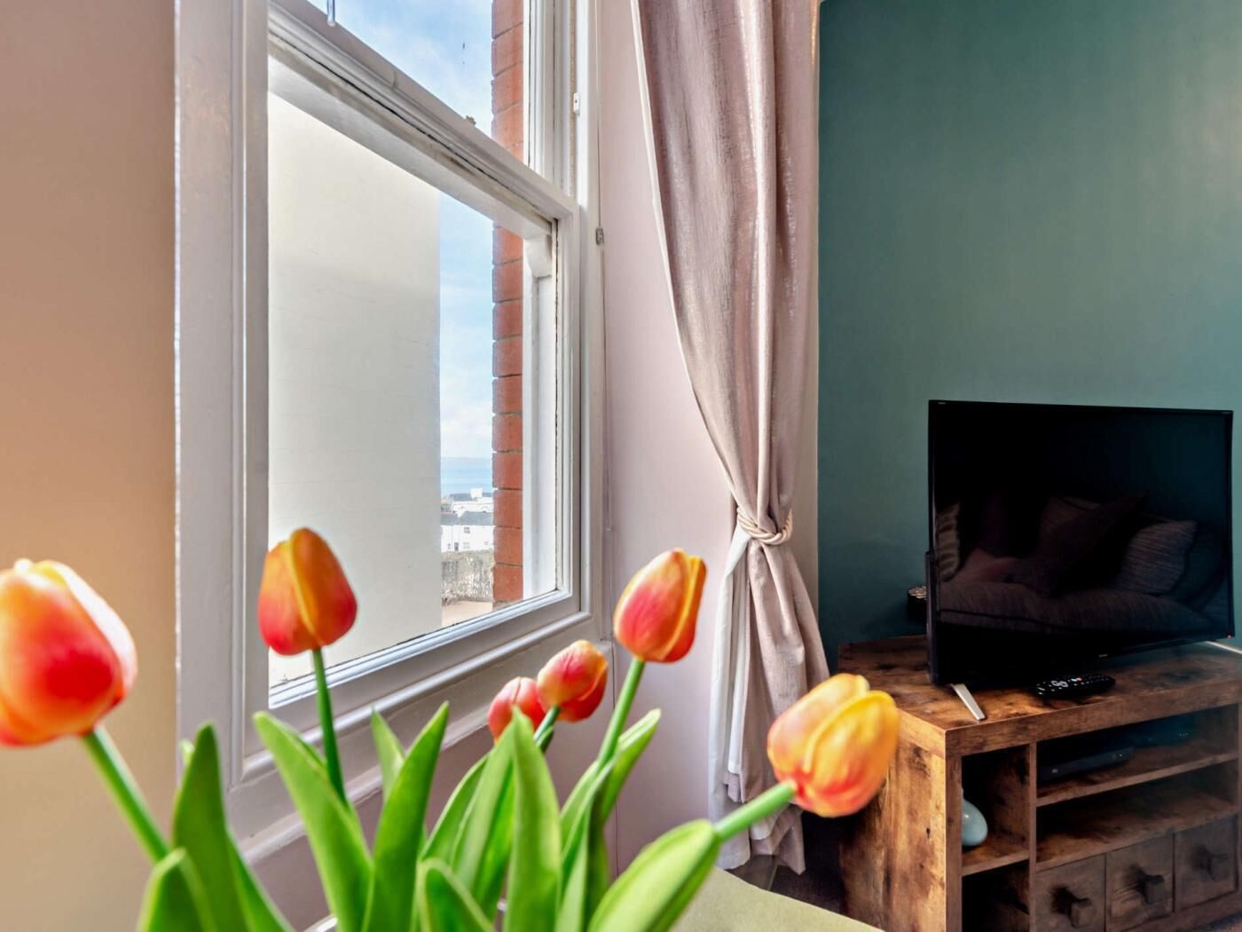 Ein Zimmer mit Fenster, einem Fernseher auf einem Holzgestell und einer Vase mit Tulpen.