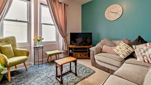 A living room with a sofa, a chair, a TV, a wooden table, a small stool, and a vase with flowers. - 2 Bed in Tenby (oc-fb122) (Tenby)