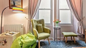 A room with a green armchair, a small side table with a vase of flowers, a lamp, and a window with curtains. - 2 Bed in Tenby (oc-fb122) (Tenby)