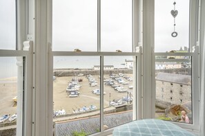 Blick aus einem Fenster auf einen Yachthafen mit angestaubten Booten, einen Sandstrand und ein Steinhaus im Hintergrund.