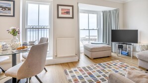A modern living room with a glass dining table, a comfortable sofa, a television, and a view of the sea. - 2 Bed in Tenby (oc-fb113) (Tenby)