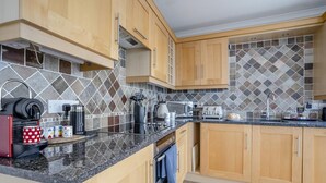 A modern kitchen with wooden cabinets, a tiled backsplash, and various appliances including a coffee machine and toaster. - 2 Bed in Tenby (oc-fb113) (Tenby)