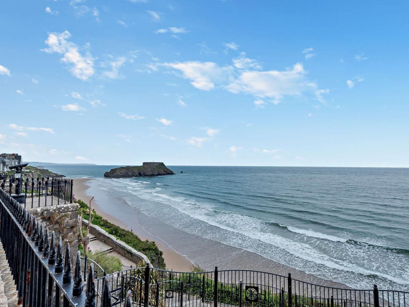 A coastal view with a rocky outcrop, a promenade, and a clear sky.