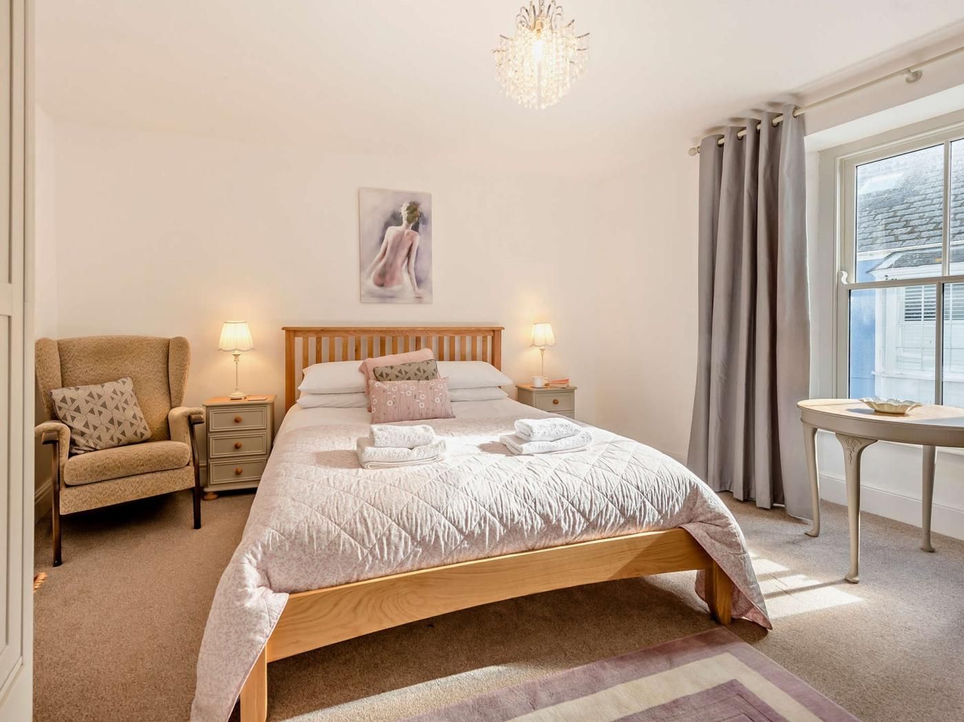 Une chambre à coucher avec un lit, une chaise, une table de chevet, une lampe, un tableau au mur et une fenêtre avec des rideaux.