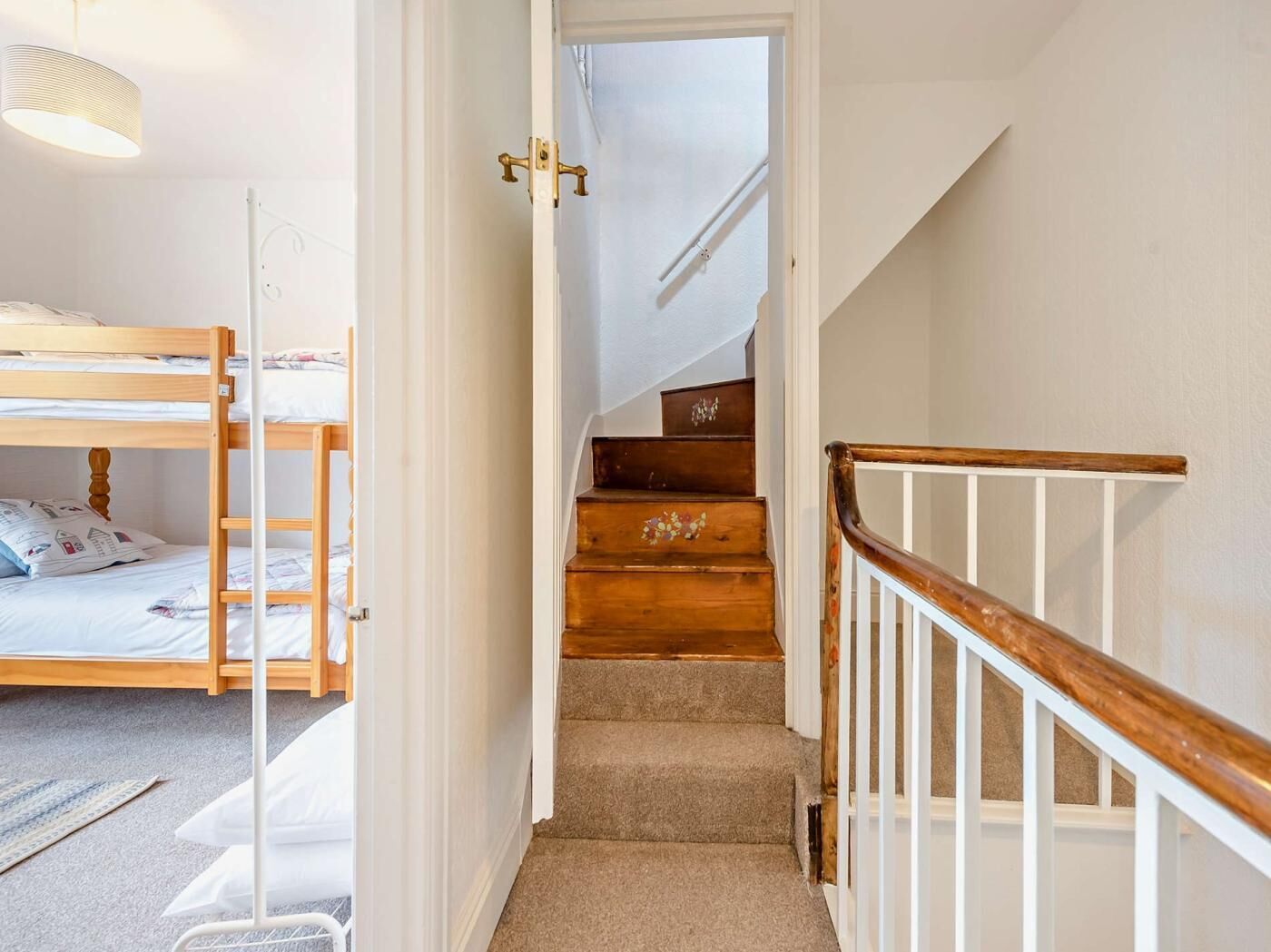 A narrow hallway with a wooden staircase leading to a landing. A bunk bed is visible on the left, and a white railing is on the right.