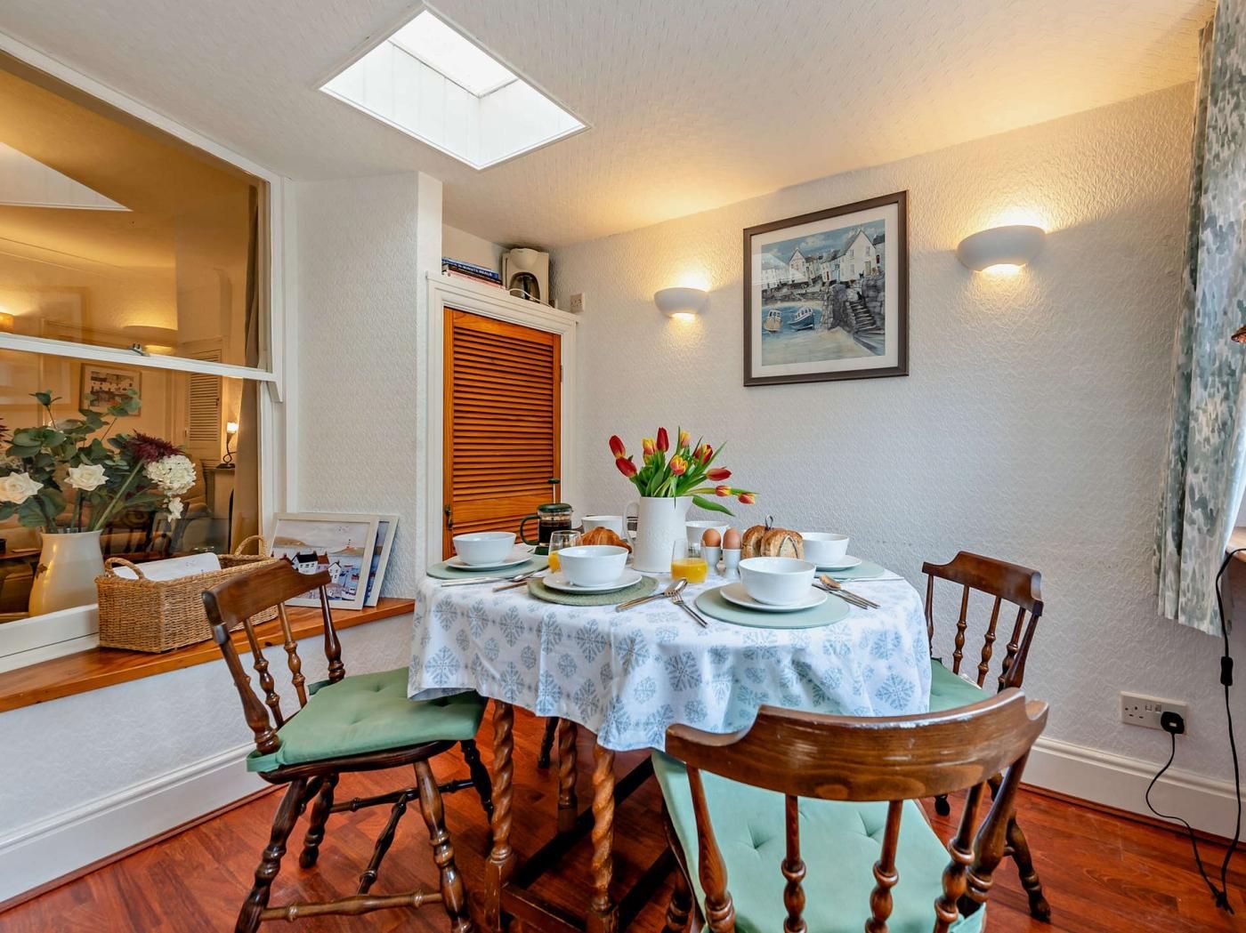 A dining area with a table set for four, chairs, a window with a view, a wall-mounted light, and a painting.