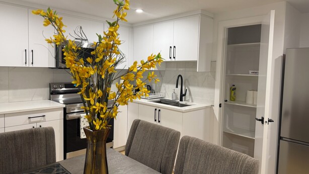 A modern kitchen with white cabinets, a stainless steel refrigerator, a dining table with grey chairs, and a vase with yellow flowers. - Spacious 2BR Suite|Near WEM|Families|Contractors (Edmonton)