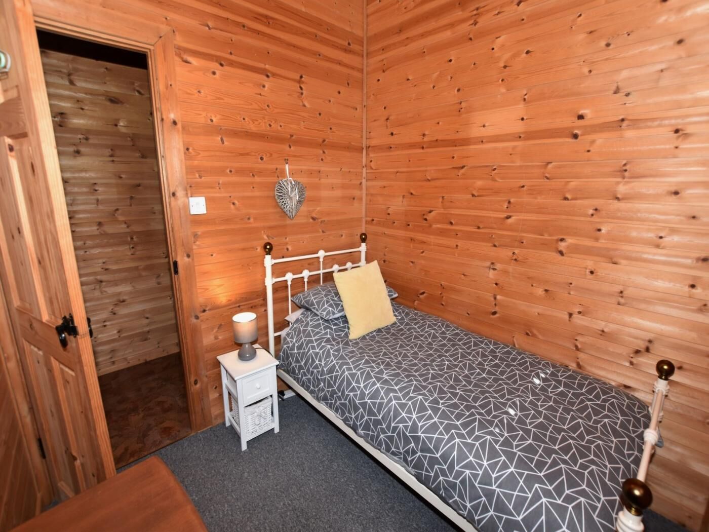 A small, single-bed room with wooden walls, a bedside table with a lamp, and a geometric-patterned bedspread.