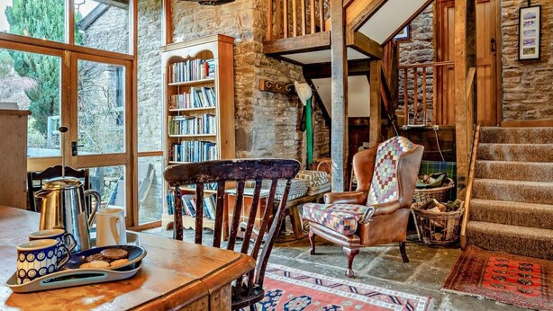 Um interior rústico com vigas de madeira aparentes, uma parede de pedra, uma estante de livros, uma poltrona de couro e uma escada.