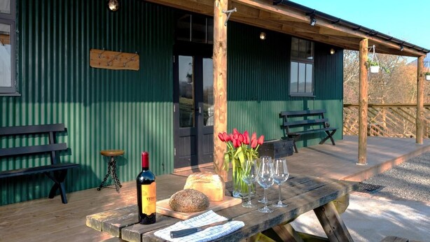 A wooden picnic table with a bottle of wine, glasses, bread, and tulips on a terrace with a green building in the background. - 2 Bed in Abergavenny (oc-bn246) (Pantygelli)