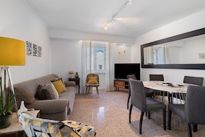 A modern living room with a sofa, a dining table with chairs, a large mirror, and a window with curtains.