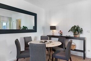 Un coin repas avec une table ronde, quatre chaises grises, un grand miroir, une lampe fixée au mur et une plante en pot.