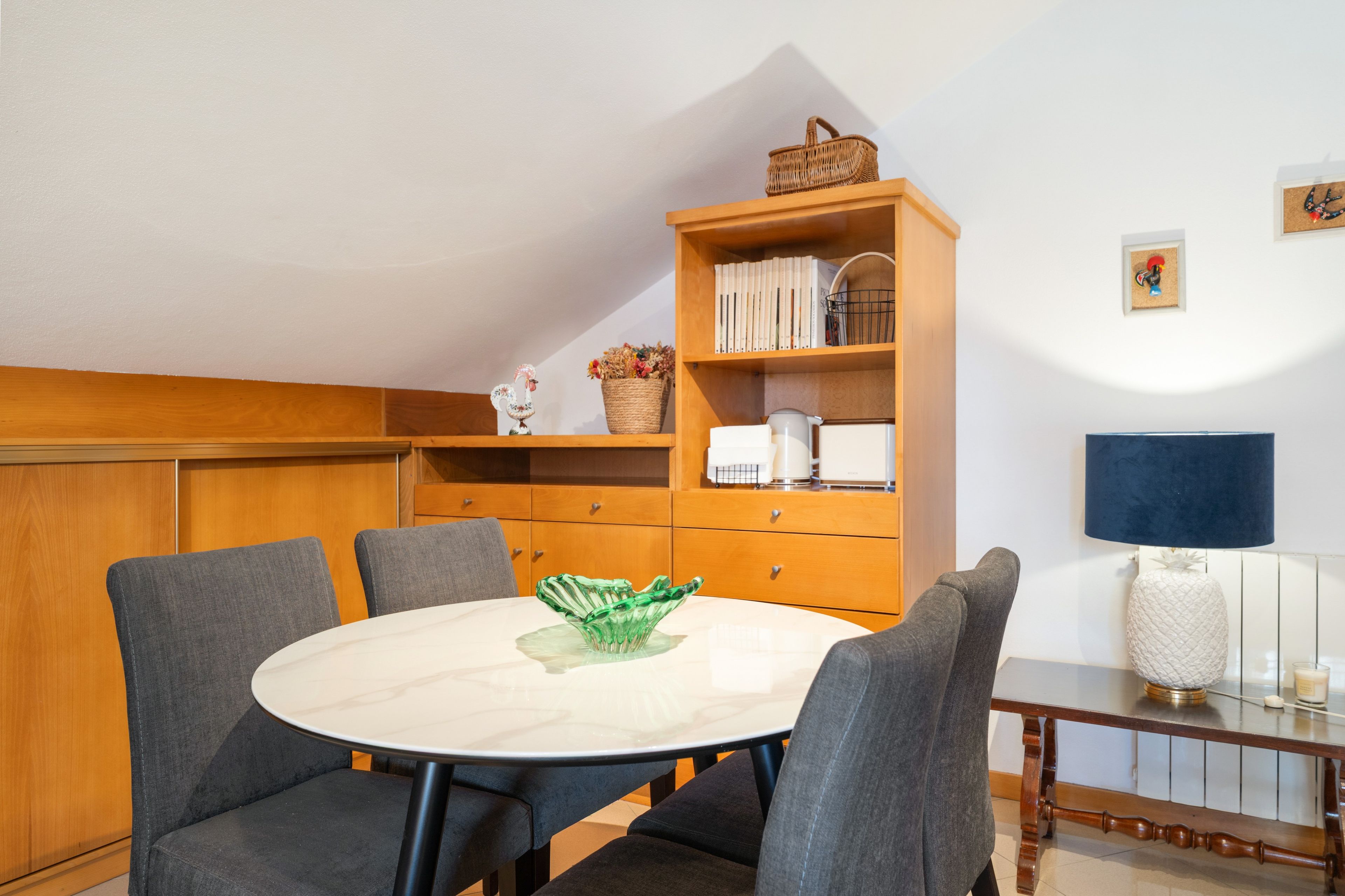 En matplats med ett runt bord, fyra grå stolar, ett träskåp med böcker och dekorativa föremål samt ett litet sidobord med en lampa.