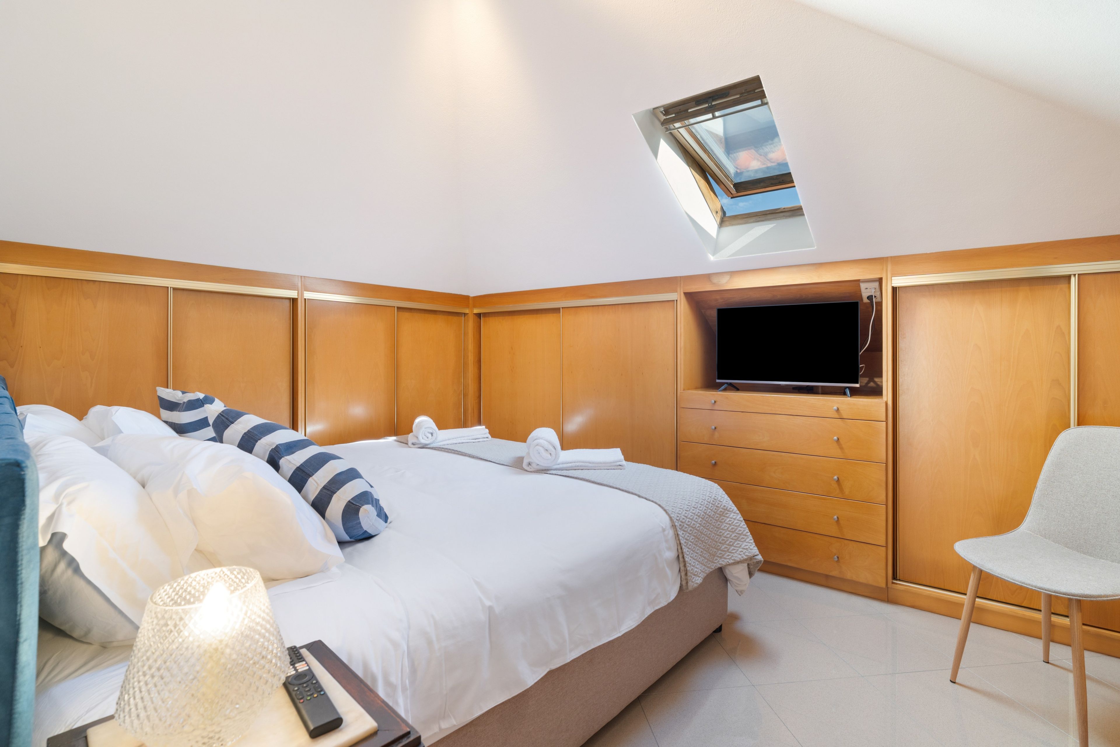 A hotel room with a bed, a television, a chair, and a skylight.