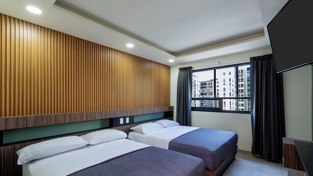 A hotel room with two beds, a wooden panel wall, a window with a view of buildings, and a flat-screen TV.