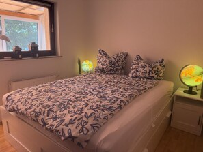 Ein Schlafzimmer mit Bett, Kissen, einem Nachttisch, einem Globus und einem Fenster mit Blick auf Bäume.