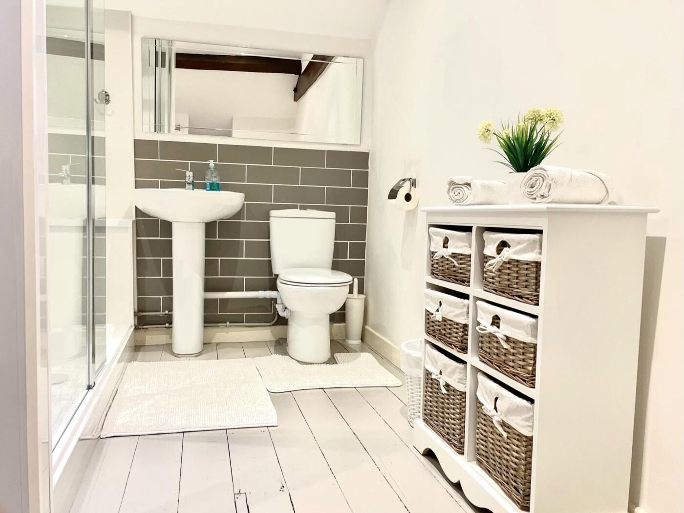 Een badkamer met een glazen douchecabine, een wit wastafelmeubel, toilet en een opbergunit met manden.