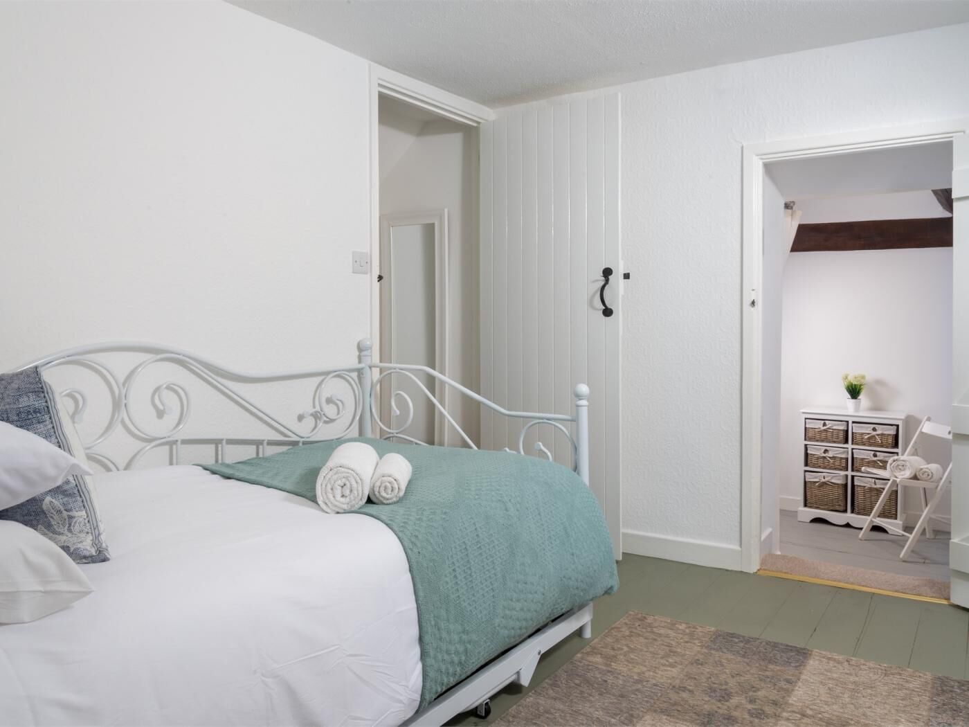 Une chambre avec un lit en métal blanc, une couverture verte et un oreiller blanc. On y trouve une commode en bois avec des paniers et une petite plante.