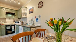 A kitchen with a dining area, including a table set for two, a clock, and a wall-mounted sign. - 1 Bed in Tenby (oc-fb125) (Tenby)