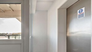 Um corredor estreito com vista para o mar através de uma janela, uma porta com um aviso e chão de azulejos.