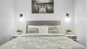 Un dormitorio con cama, cabecero, mesitas de noche, dos luces fijadas en la pared, un cuadro de un muelle y dos plantas en macetas.