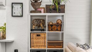 Una sala de estar con un sofá, una estantería con objetos decorativos, un cuadro enmarcado, una planta y un extintor.