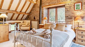 Um quarto com parede de pedra, teto de madeira, cama com roupa de cama branca, uma cadeira e uma mesa de cabeceira com abajur.